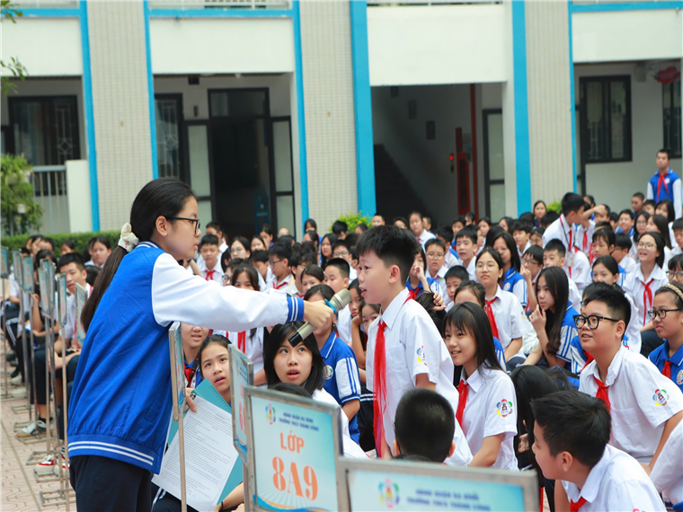 A person holding a microphone in front of a group of students

Description automatically generated