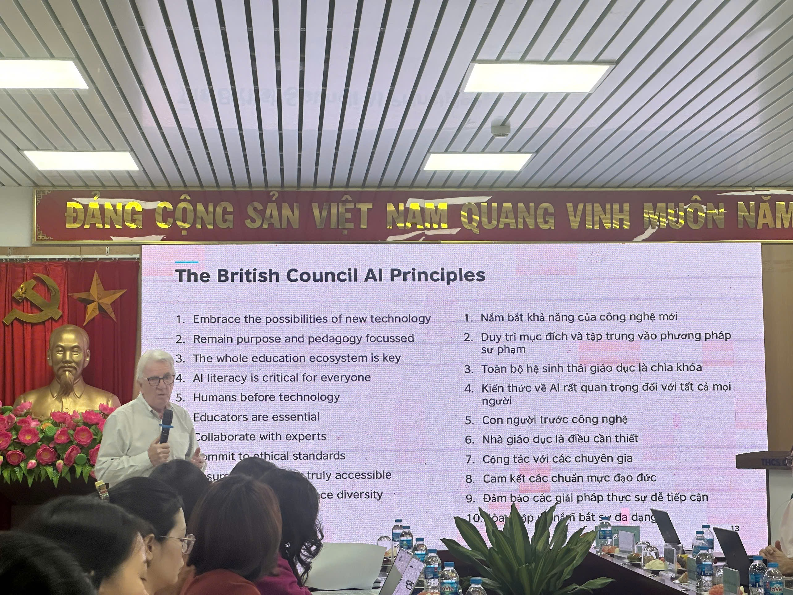 A person speaking into a microphone in front of a large white board with text

Description automatically generated