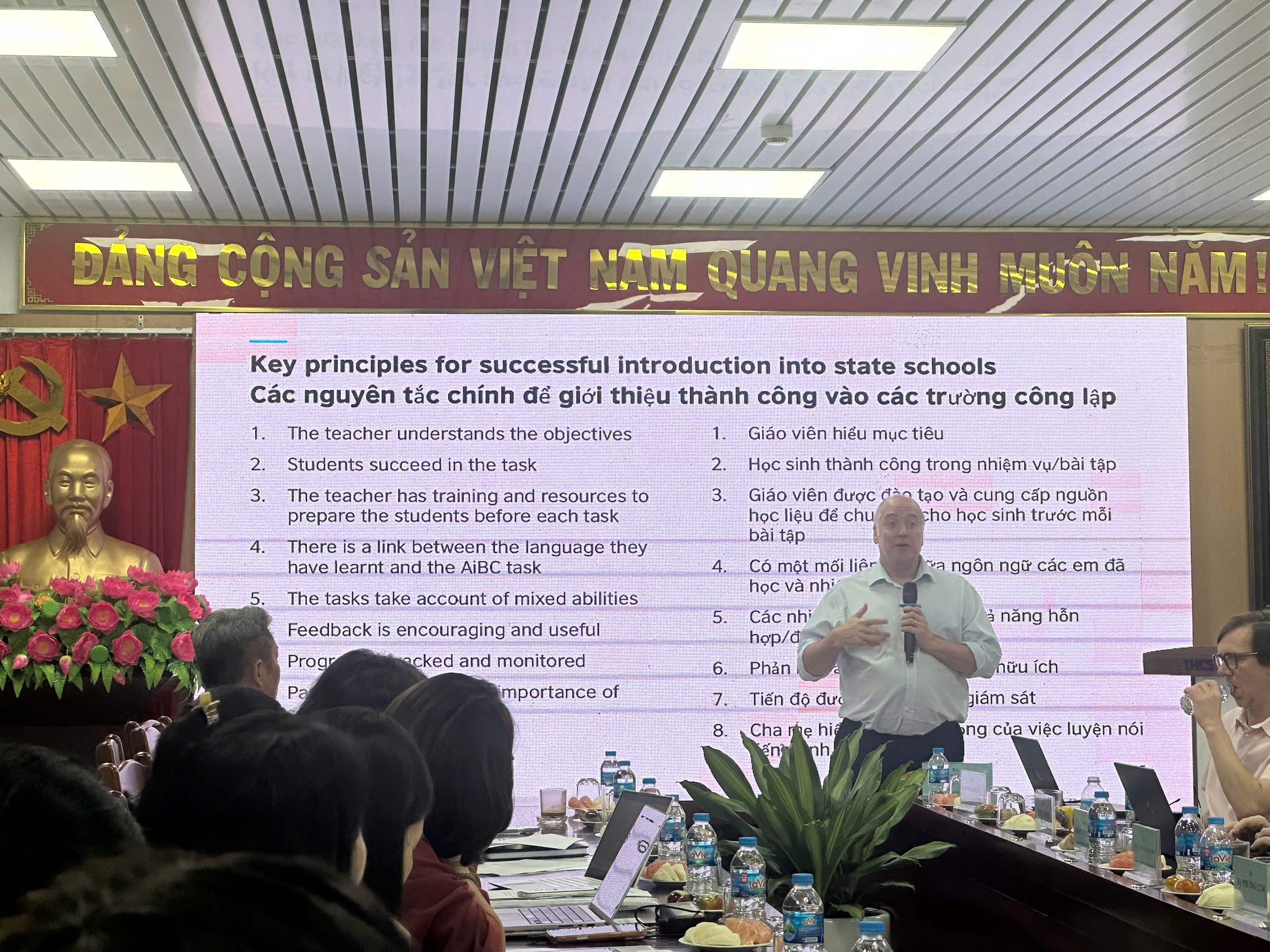 A person standing in front of a large screen with a group of people

Description automatically generated